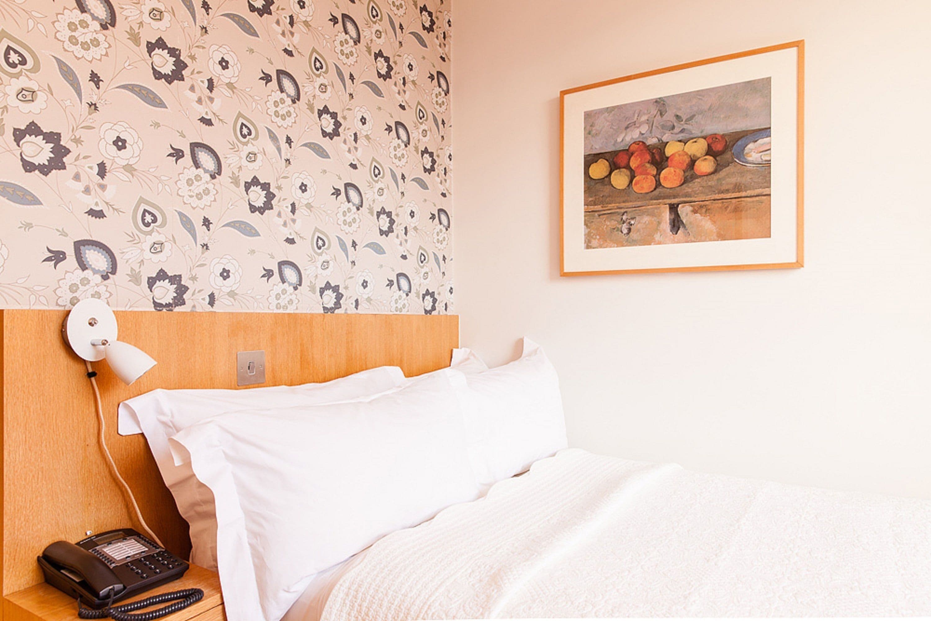 A hotel room with a patterned wallpaper, a framed painting of fruit, a bedside telephone, and a neatly made bed.