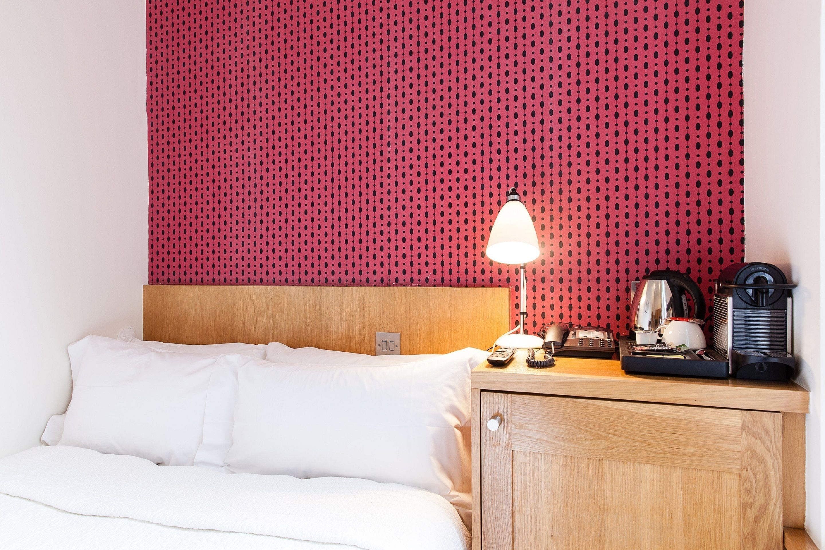 Een net opgemaakt bed met witte lakens, een houten hoofdbord, een rode muur met stippen, een bedlampje, een klein houten nachtkastje met een waterkoker en een koffiezetapparaat.