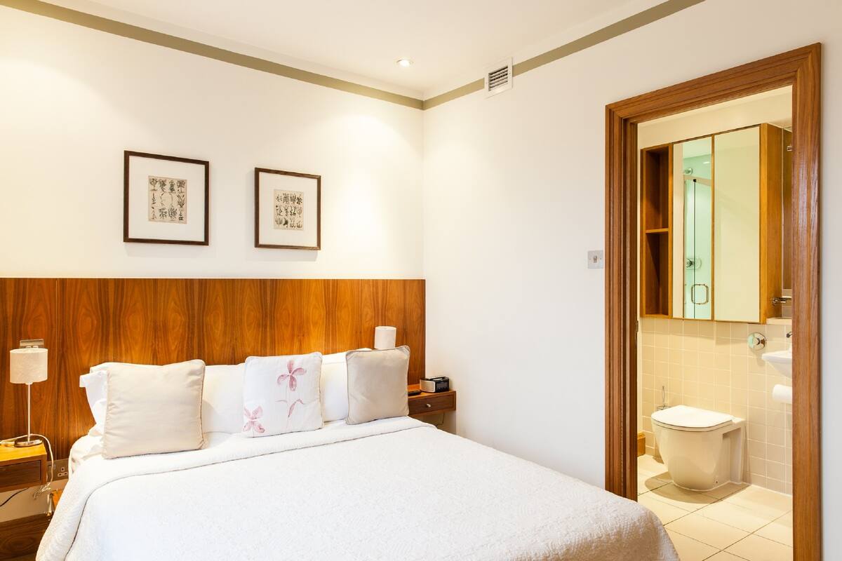 A hotel room with a wooden headboard, a bed with white bedding, two framed pictures on the wall, a bedside table with a lamp, and a view of the bathroom through an open door.