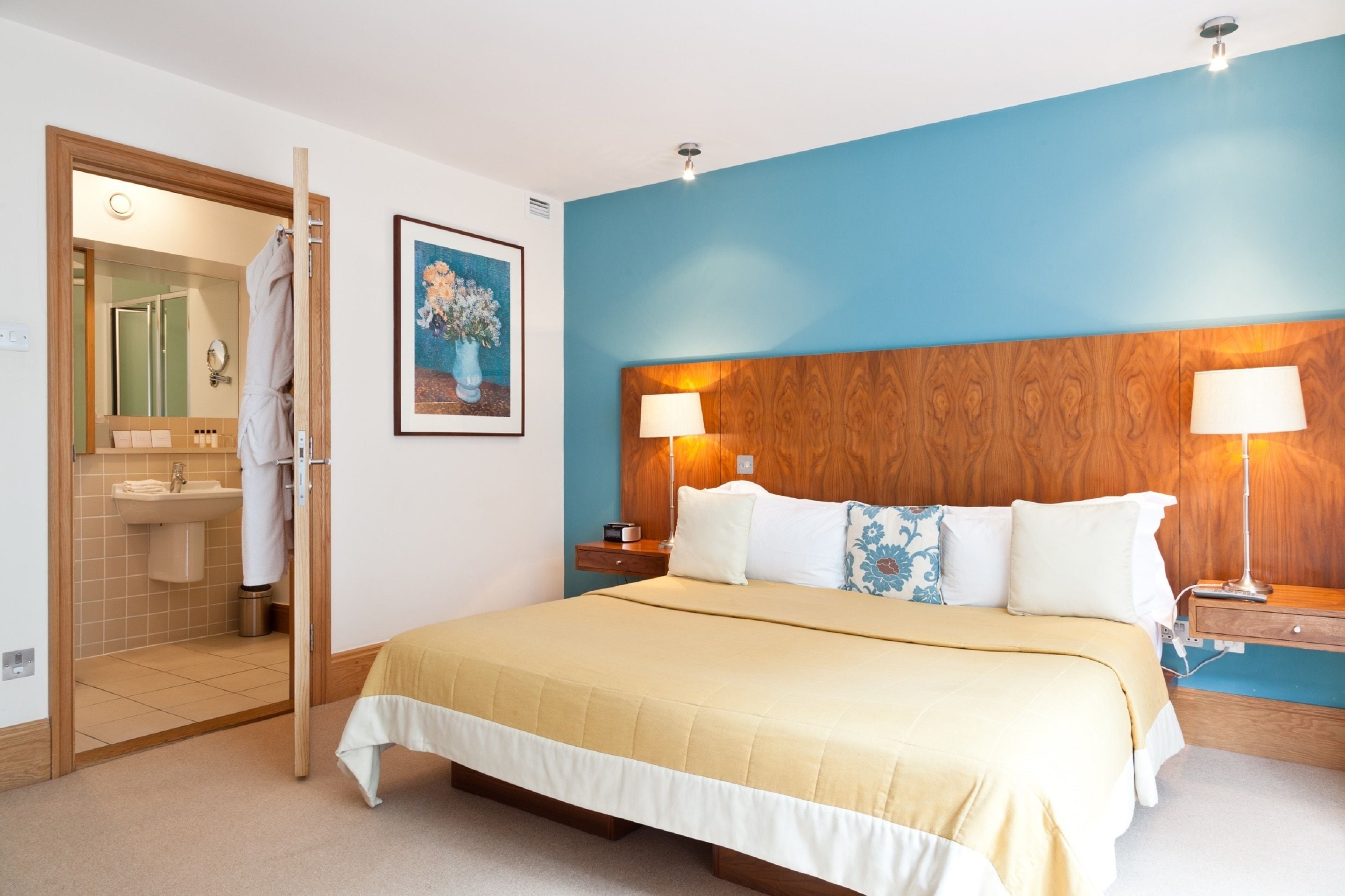 A bedroom with a bed, bedside lamps, a wooden headboard, a painting, and a bathroom visible through an open door.