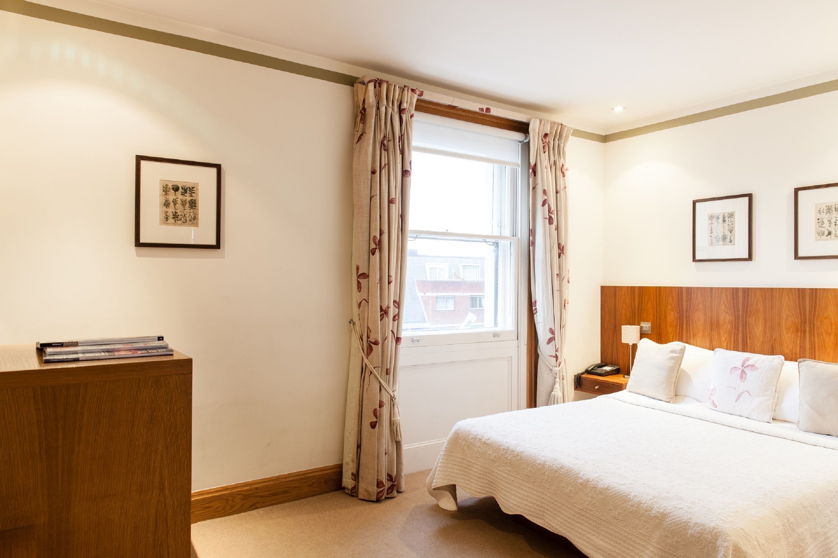 A hotel room with a bed, wooden headboard, a nightstand, a bedside table, a window with curtains, and framed artwork on the walls.