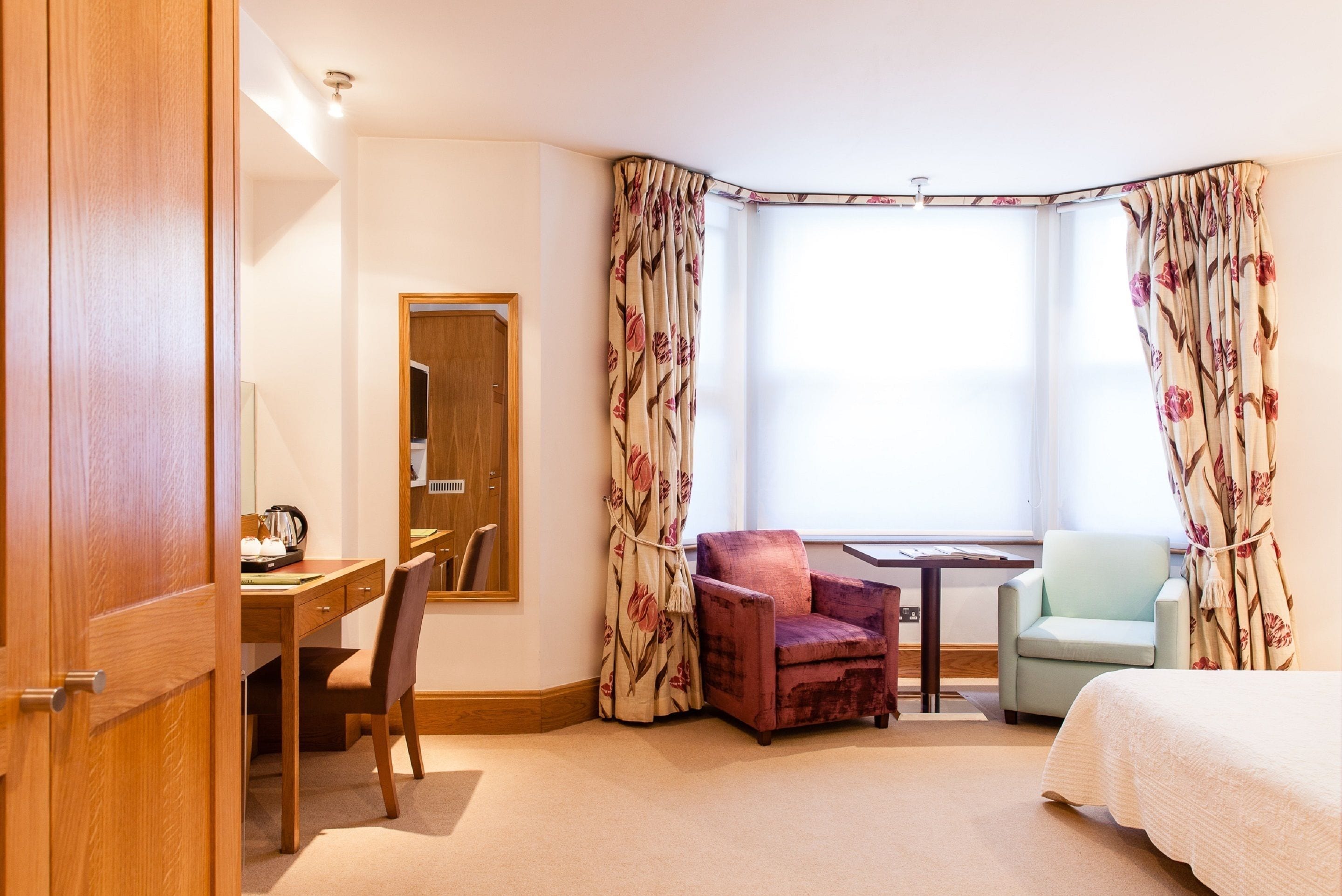 A hotel room with a wooden desk, two chairs, a bed, and a large window with floral curtains.