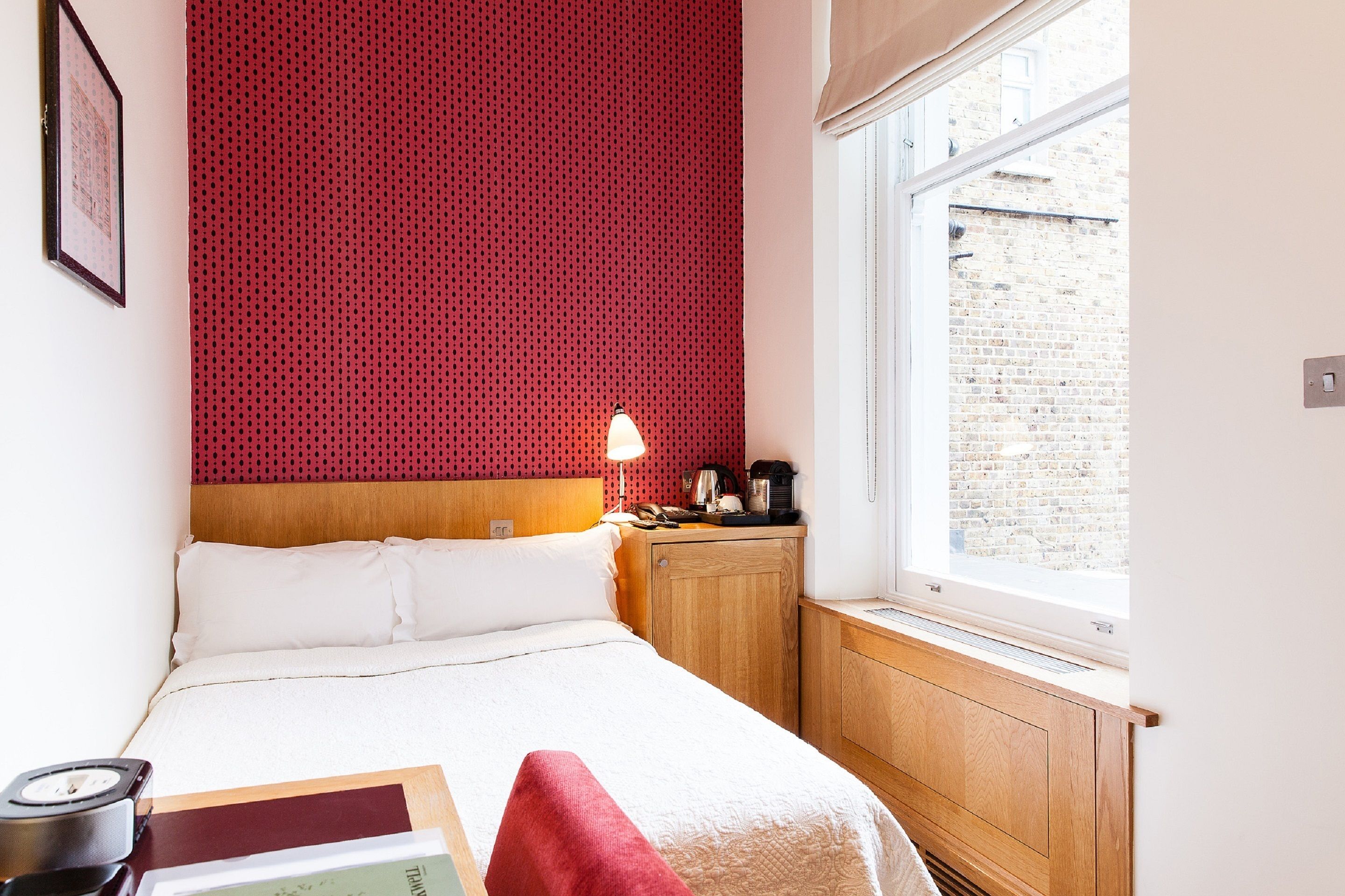 A neatly arranged hotel room with a bed, a bedside table with a lamp, a small cabinet, and a window with a view of a brick building.
