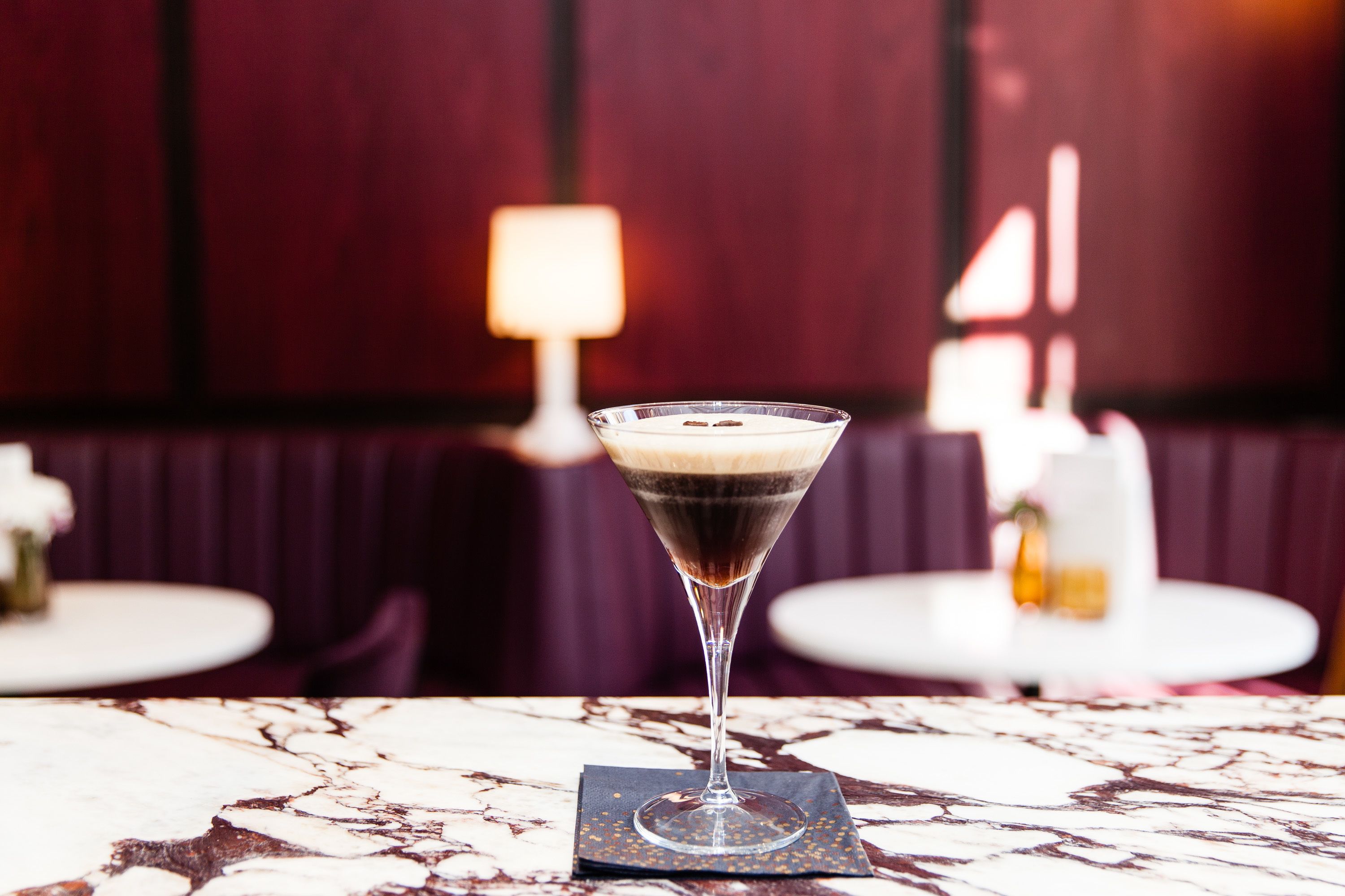 A martini glass on a marble table with a napkin underneath, set in a dimly lit room with a purple wall and a small table with a candle in the background.