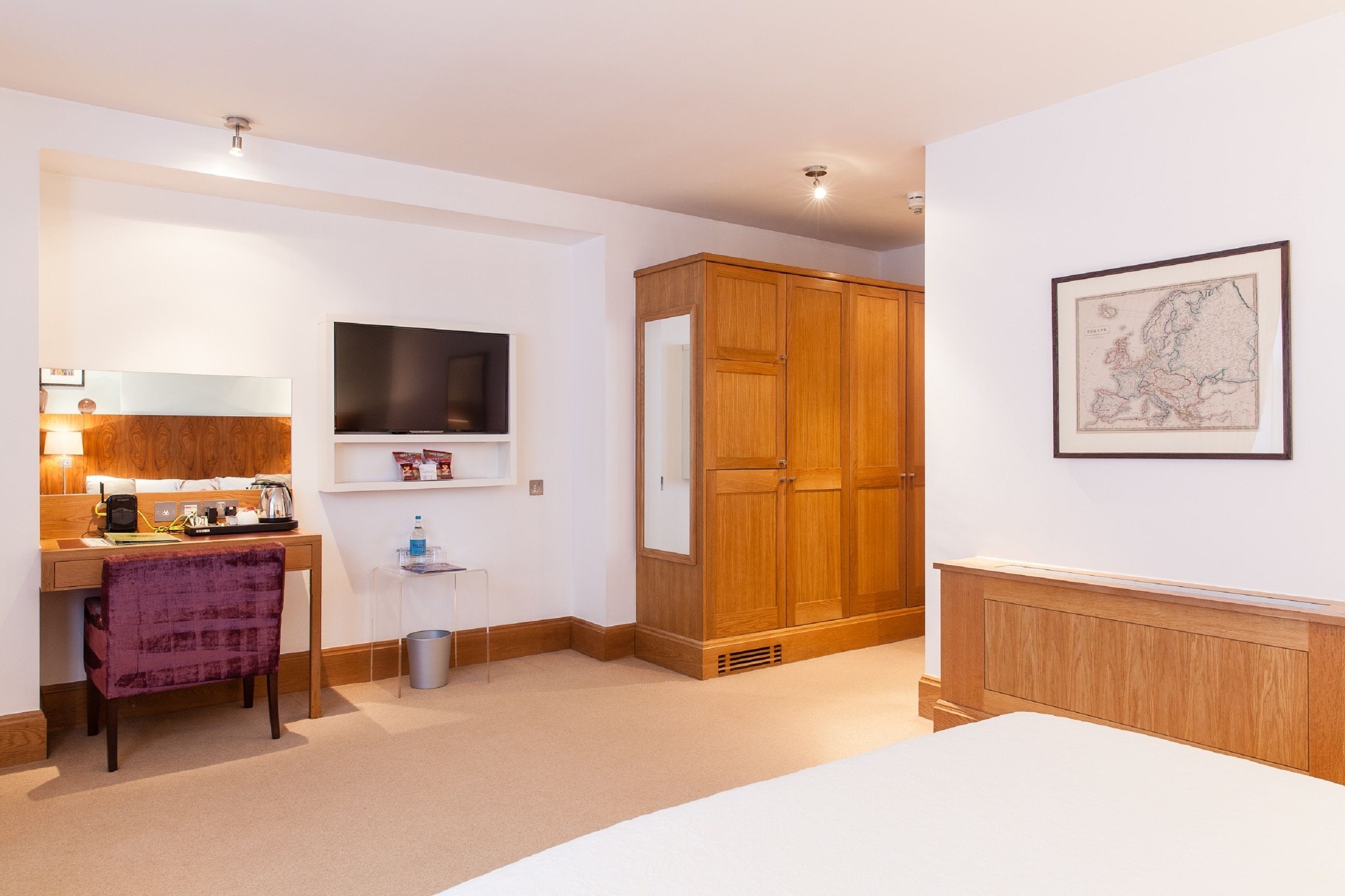 A hotel room with a wooden wardrobe, a television, a desk with a chair, and a framed map on the wall.