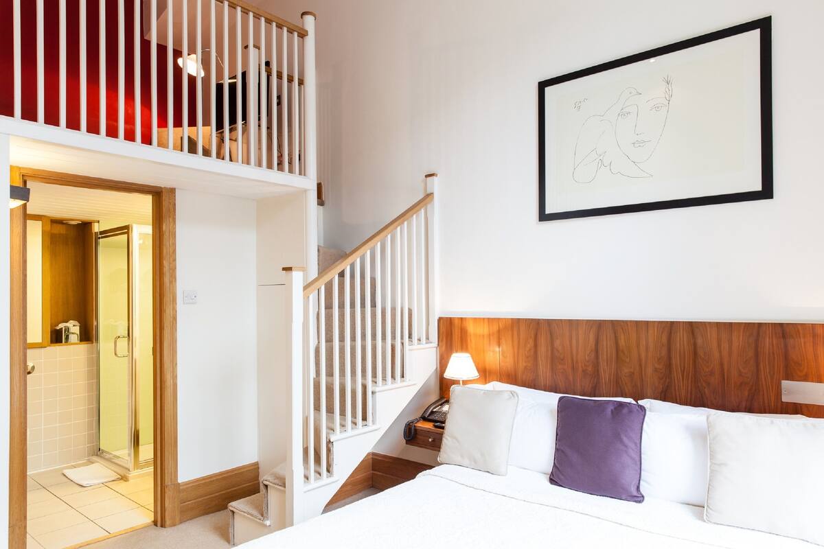 A hotel room with a wooden headboard, a bed with white and purple pillows, a nightstand with a lamp, and a framed artwork on the wall.