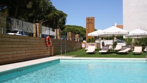 A swimming pool with lounge chairs and umbrellas, a wooden fence, and a hotel sign in the background. - Vime Islantilla (Isla Cristina)
