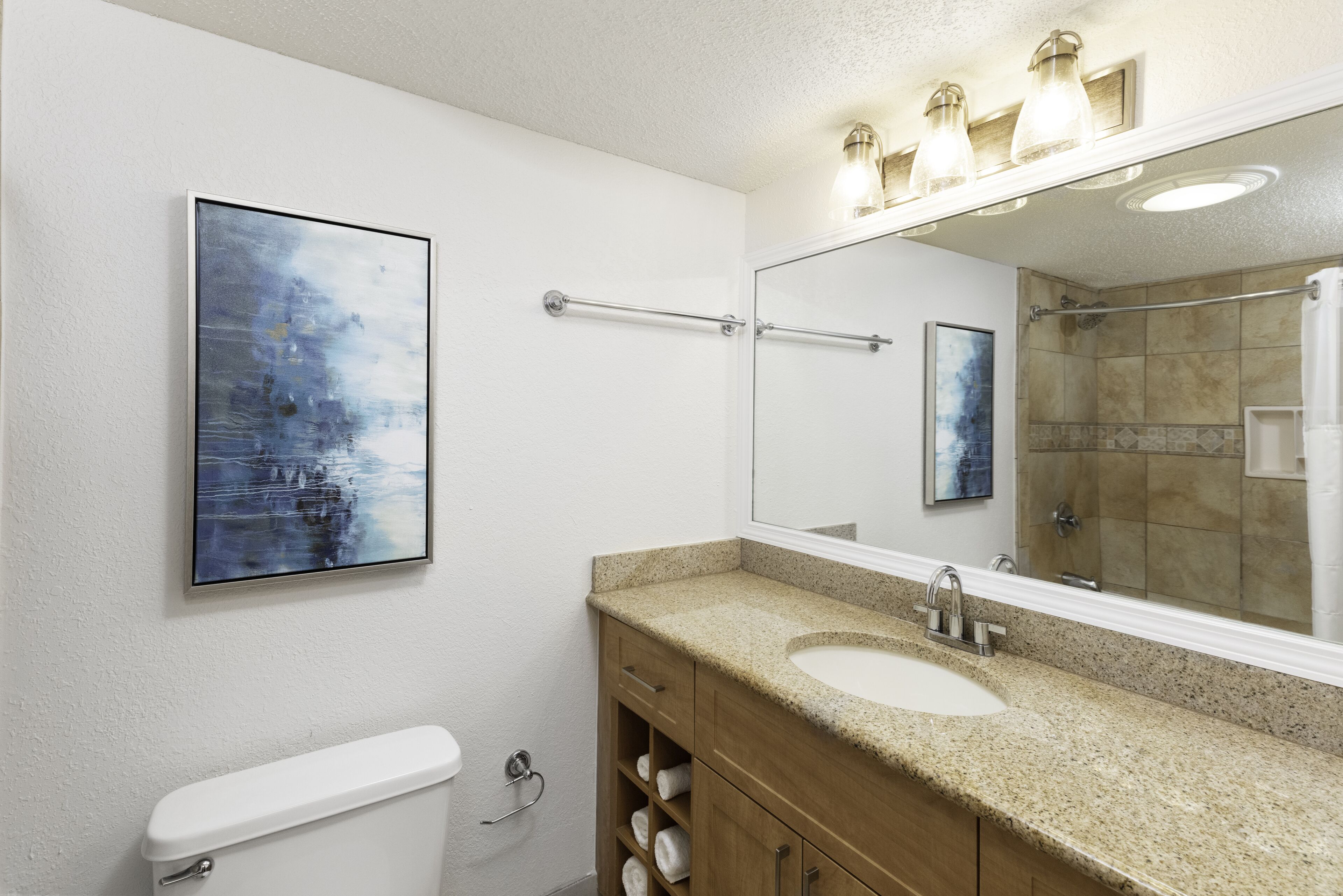 A bathroom with a white toilet, a beige countertop sink, a wooden vanity, a large mirror, and a towel rack.