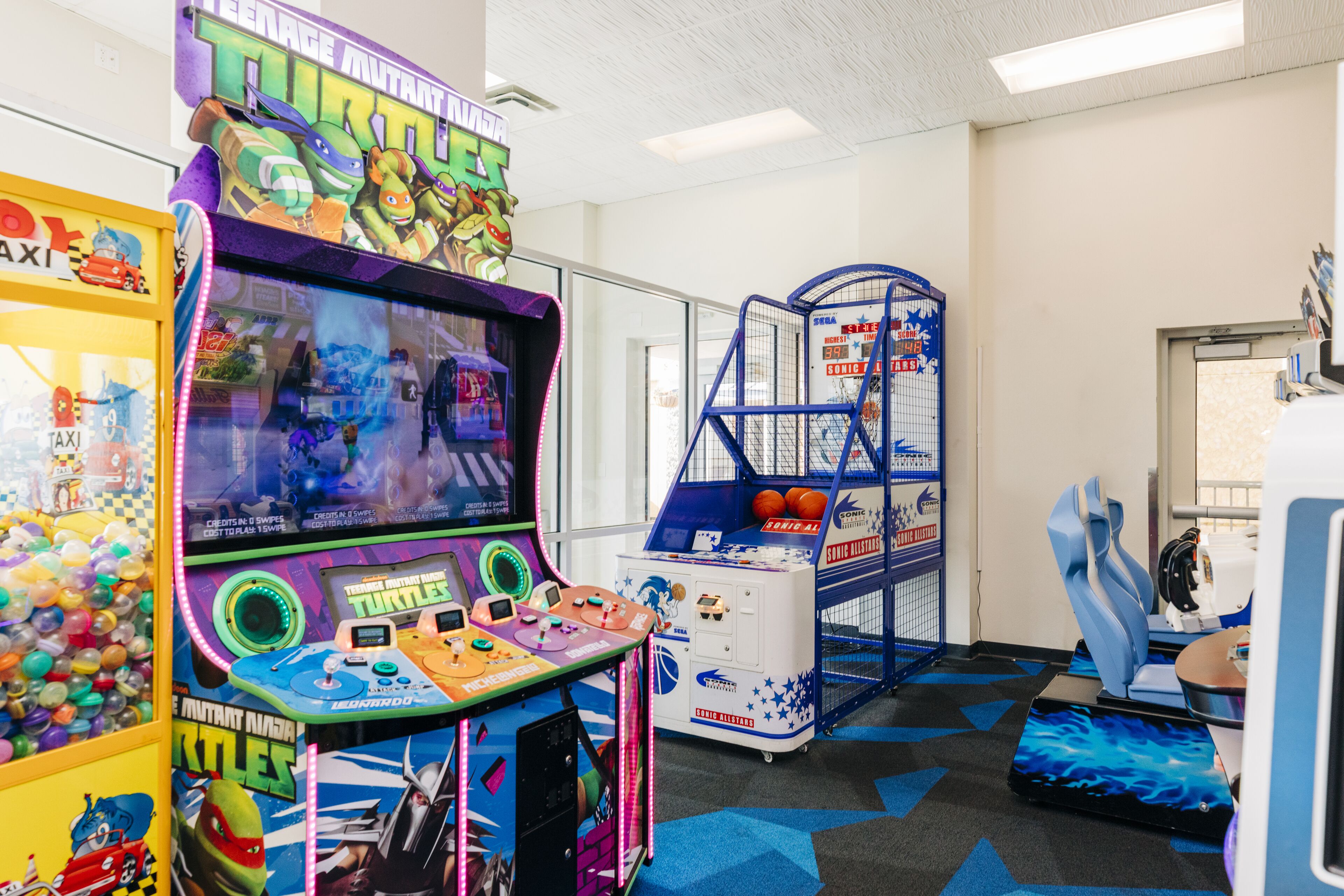 Zona de juegos recreativos con varias máquinas, incluyendo un juego de 'Teenage Mutant Ninja Turtles', y una zona de estar con sillas azules.