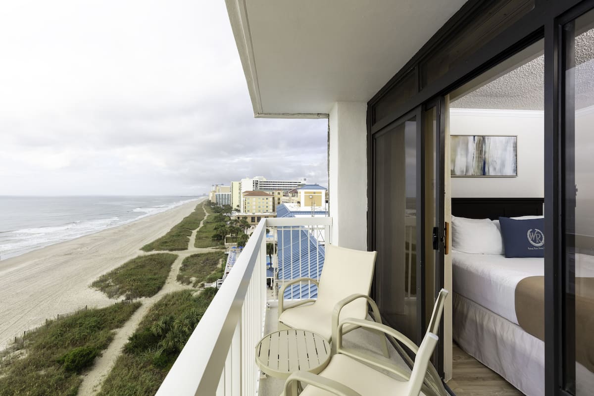 A balcony with a view of the beach and ocean. There is a bed with white bedding and a blue pillow.