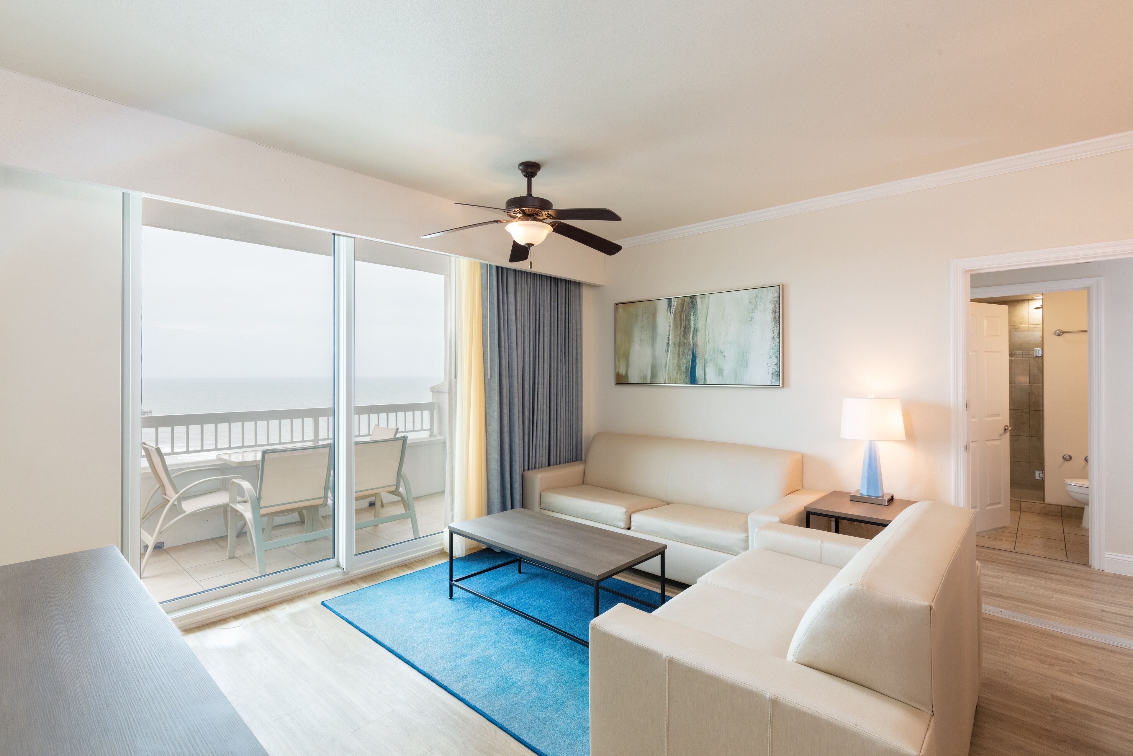 A modern hotel room with a balcony, a sofa, a coffee table, and a ceiling fan.