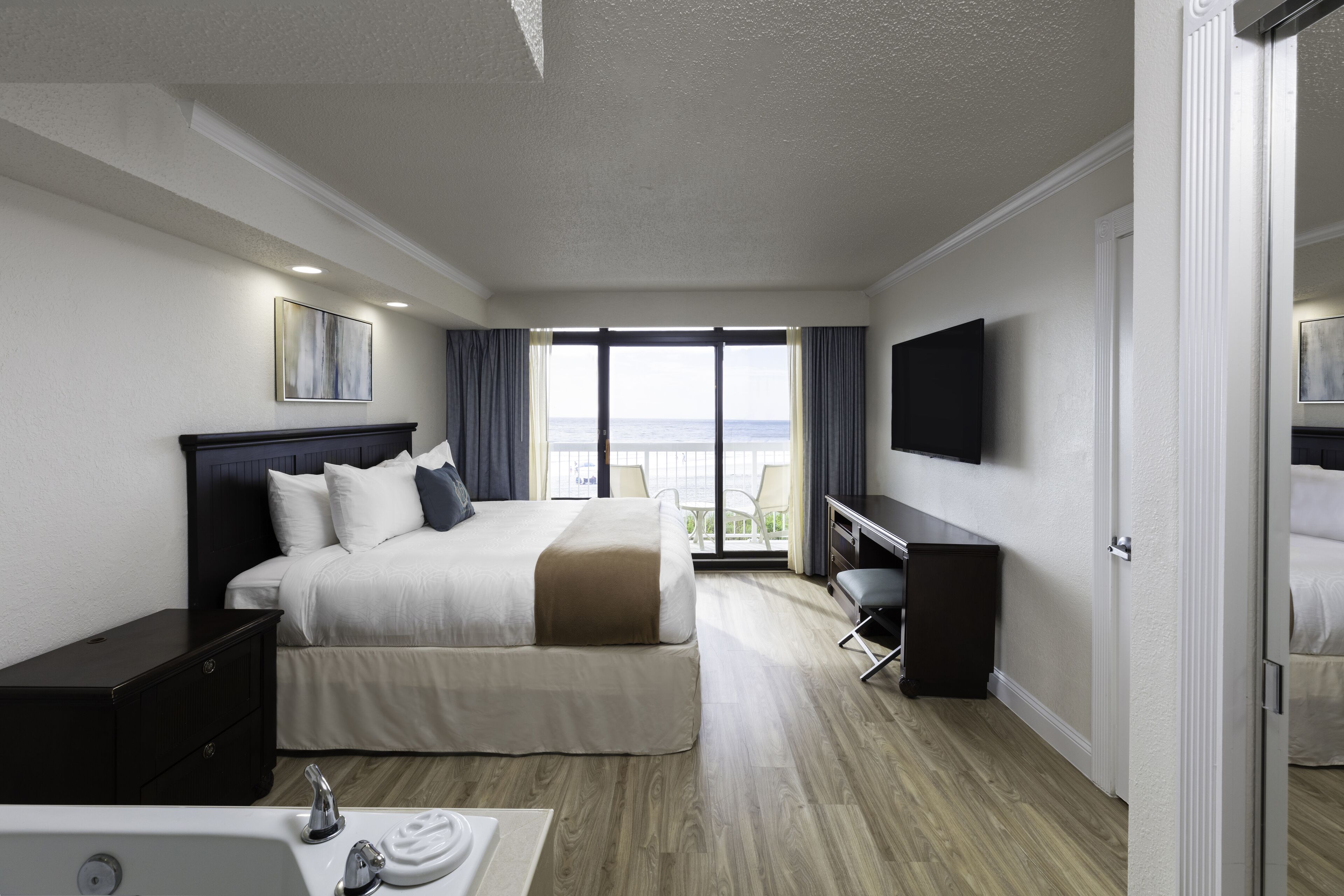 A hotel room with a bed, a desk, a TV, and a balcony with a view of the sea.