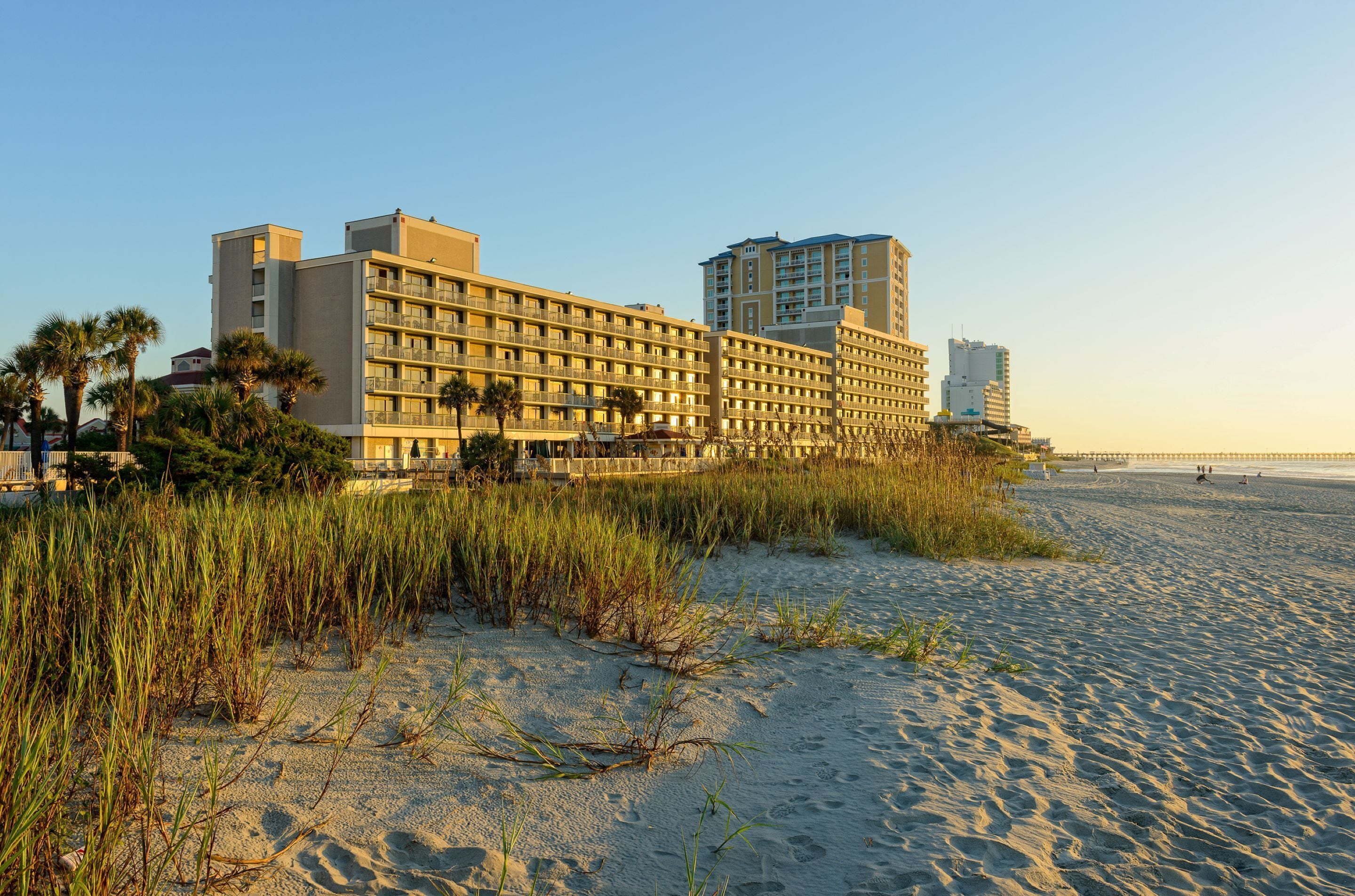 A hotel with multiple floors, a beach in the background, and a sandy foreground with tall grasses.