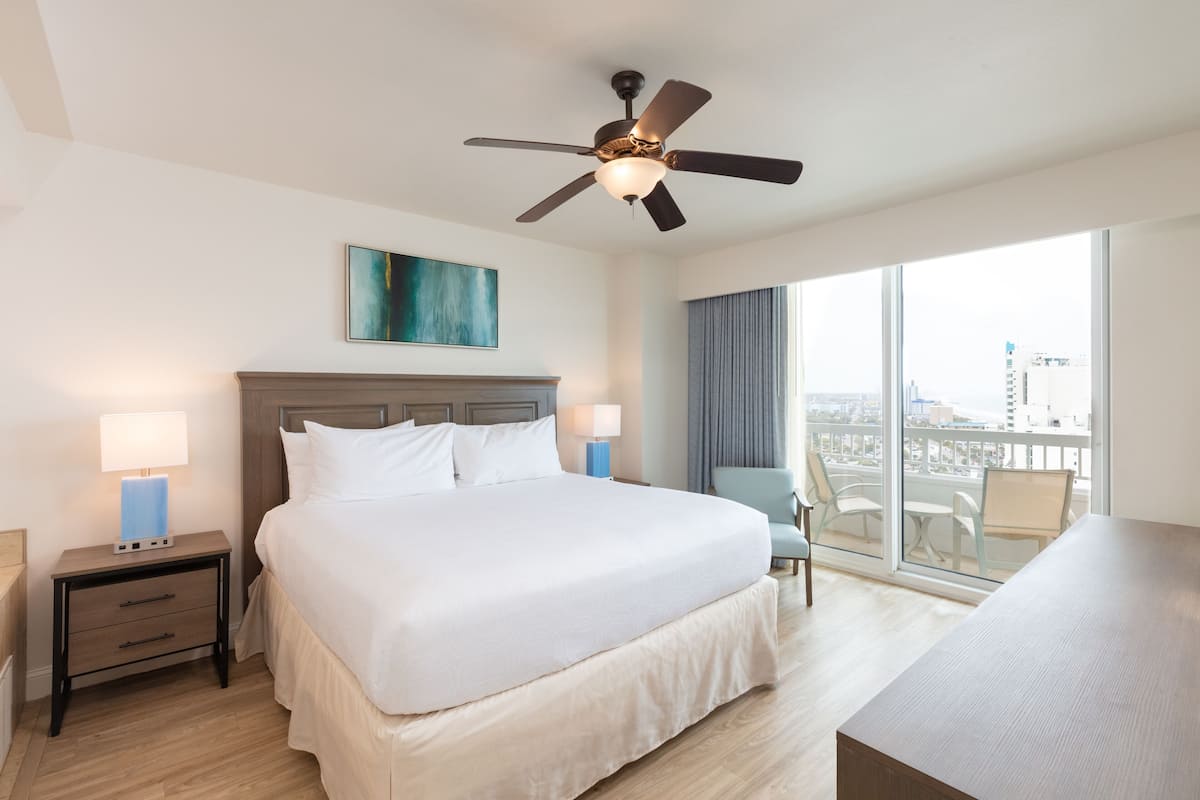 A hotel room with a large bed, a ceiling fan, a city view, and a desk with a chair.