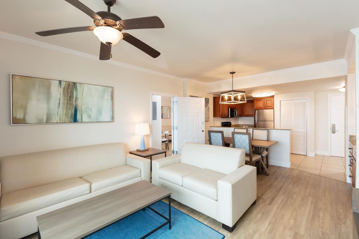 A modern living room with a sofa, coffee table, and a ceiling fan. A kitchen is visible in the background with wooden cabinets and a dining area.