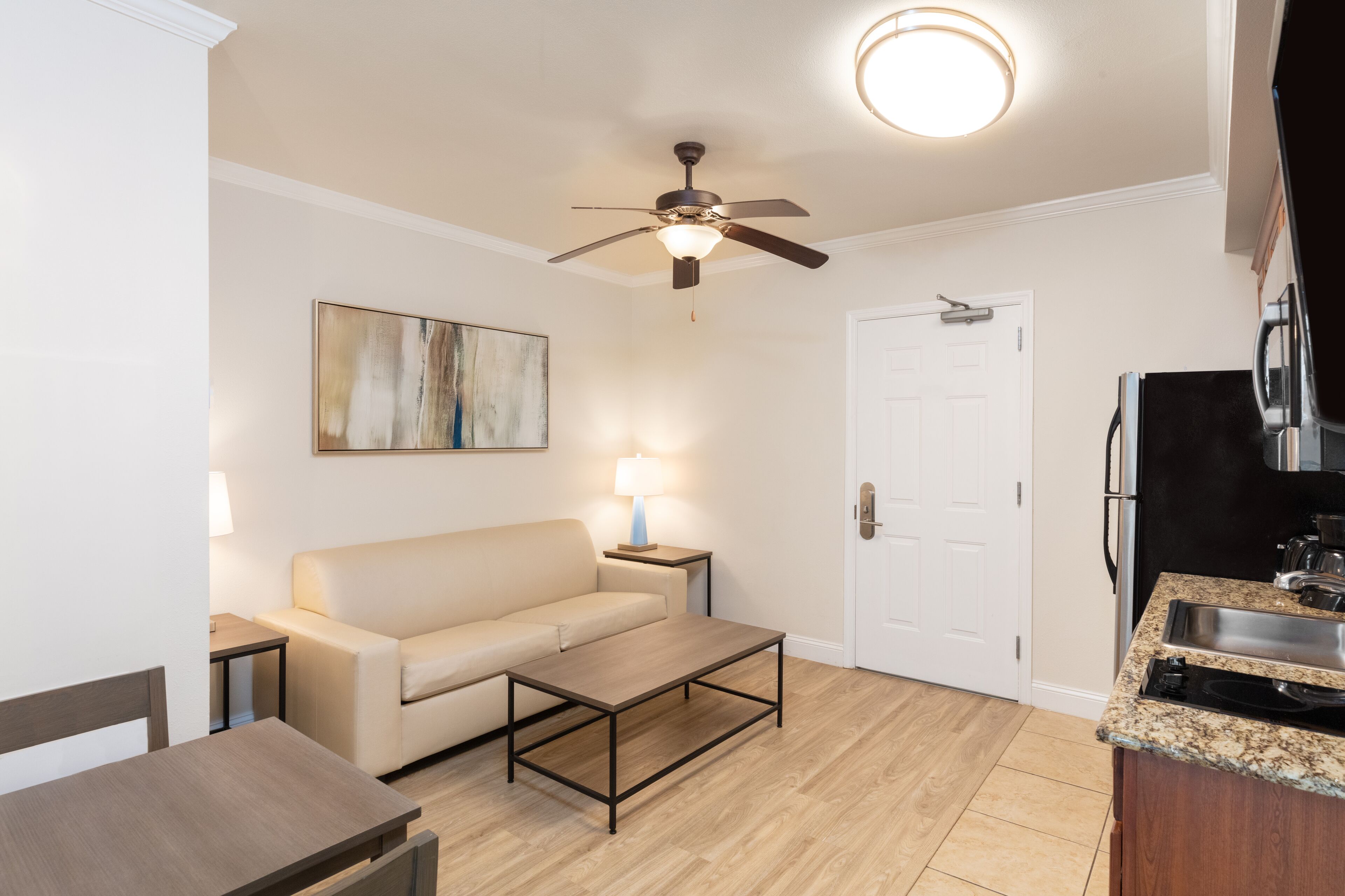 A modern living room with a beige sofa, a coffee table, a ceiling fan, and a kitchen area with a sink and stove.