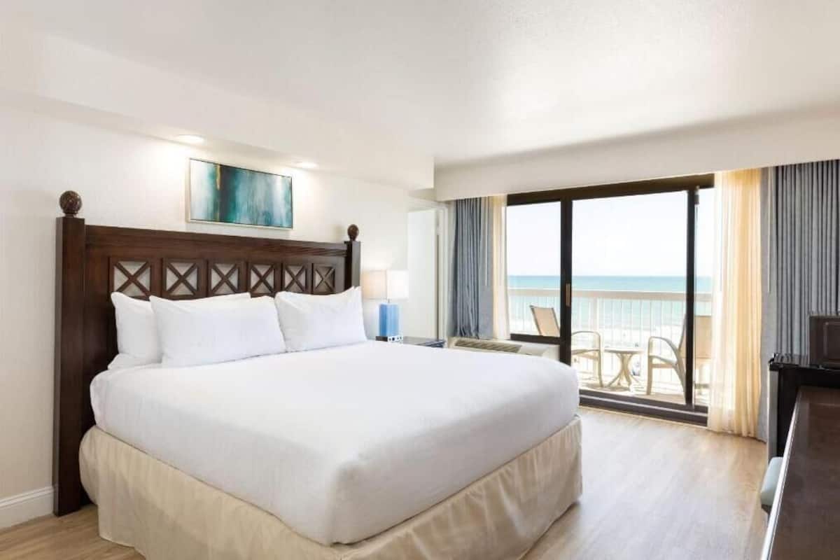A hotel room with a large bed, a painting above the bed, a view of the ocean through the balcony, and a desk with a lamp.