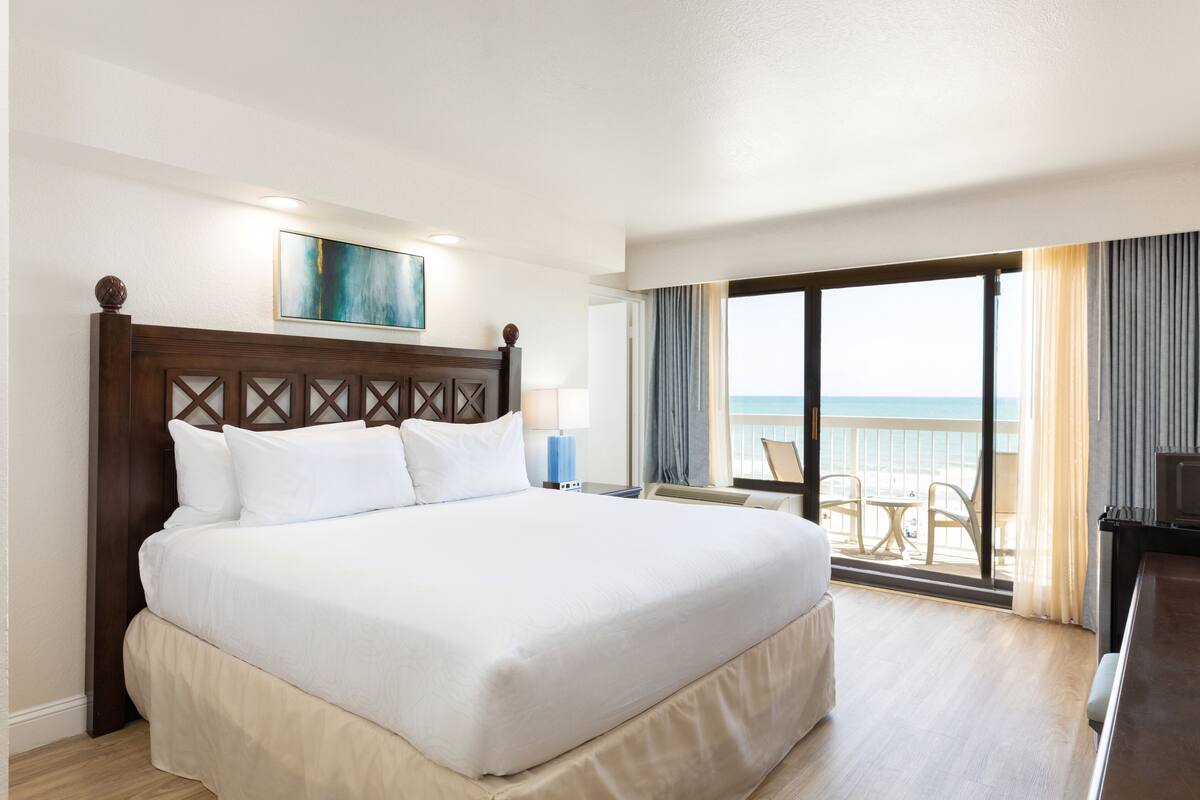 A hotel room with a large bed, a view of the ocean, and a balcony with chairs.