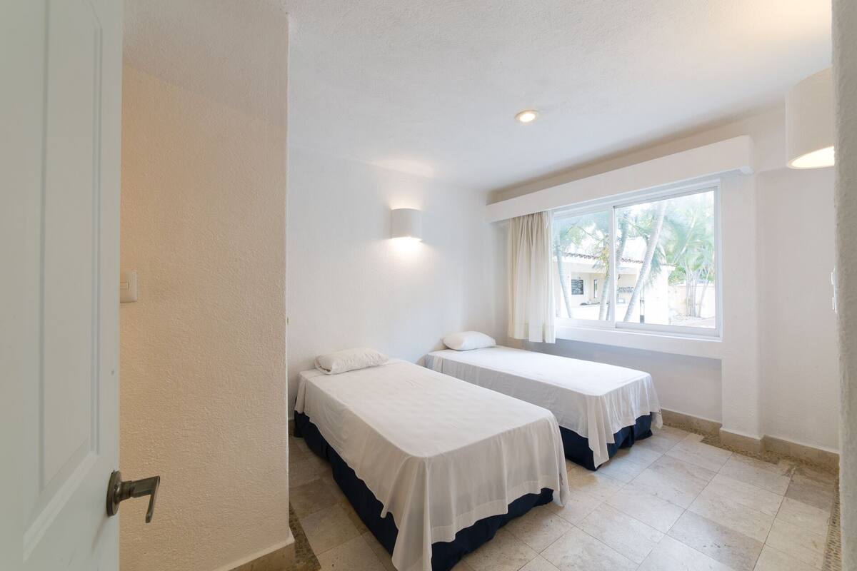 A hotel room with two single beds, a window with curtains, and a view of palm trees outside.