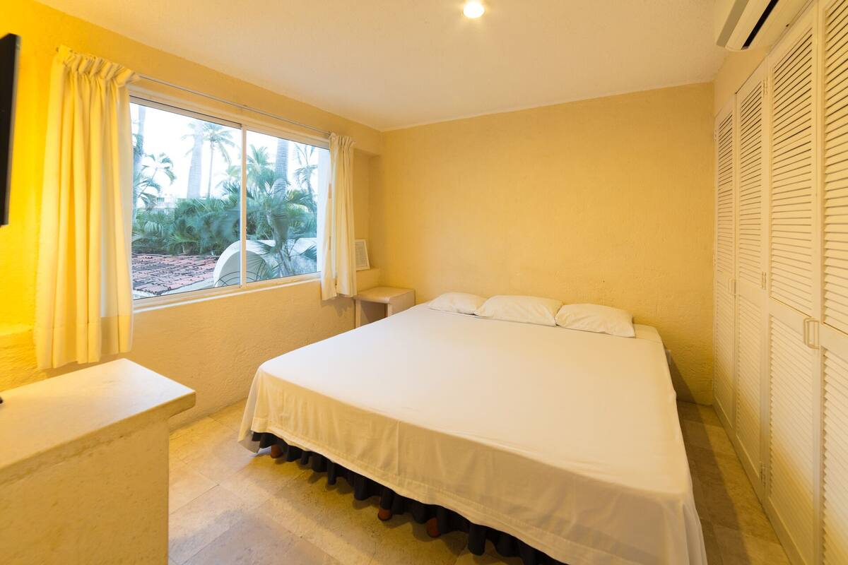 A bedroom with a bed, a nightstand, a window with curtains, and a view of palm trees.