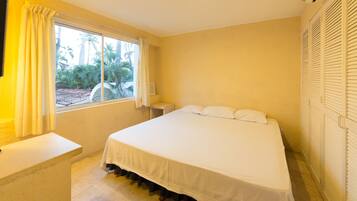 A bedroom with a bed, a nightstand, a window with curtains, and a view of palm trees.