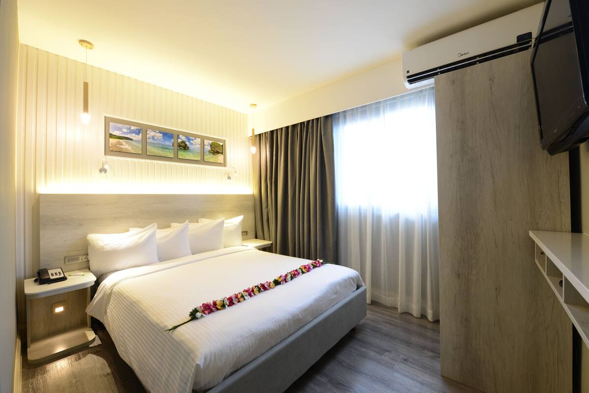 Ein Hotelzimmer mit einem großen Bett, einem Nachttisch mit Telefon, einer Wand-Klimaanlage und einem Fenster mit Vorhängen.