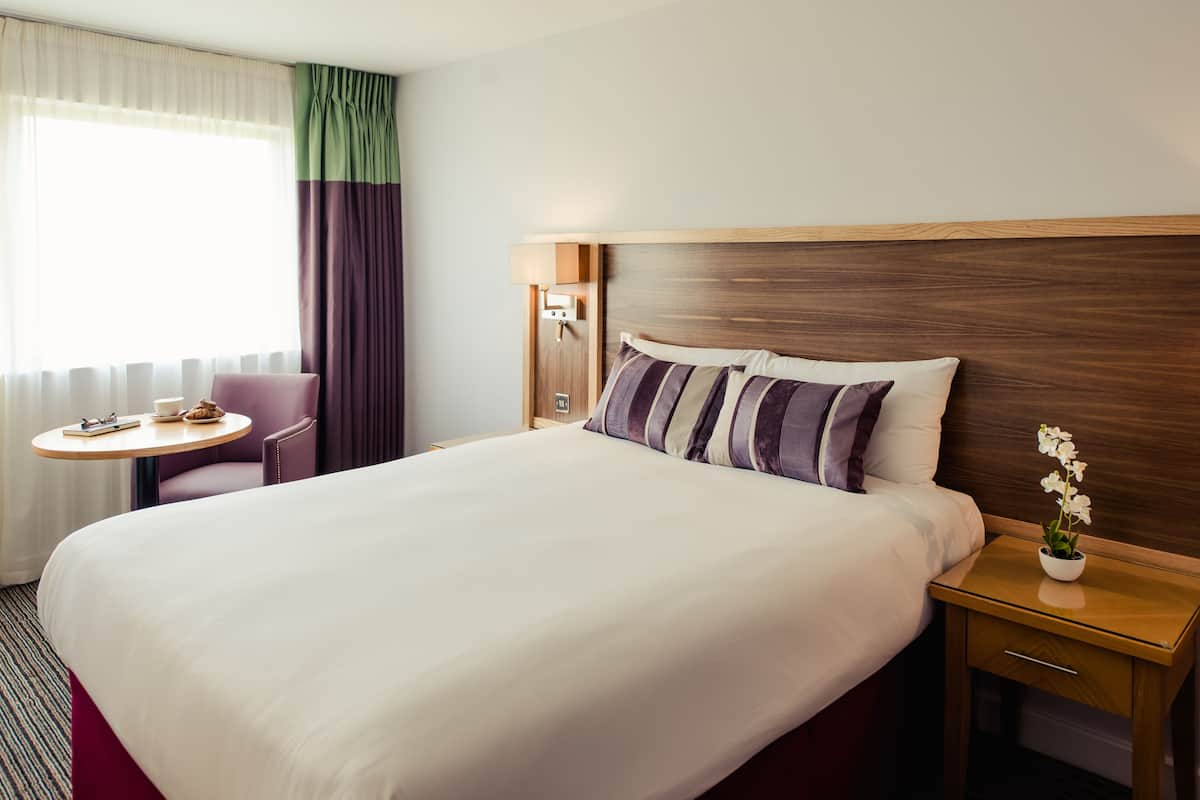 Une chambre d’hôtel avec un grand lit, une table de chevet avec une lampe, une petite table ronde avec une chaise, et une fenêtre avec des rideaux.