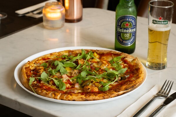 Une pizza sur une assiette blanche, avec une fourchette et un couteau à côté, un verre de bière et une bouteille de bière Heineken sur une table.