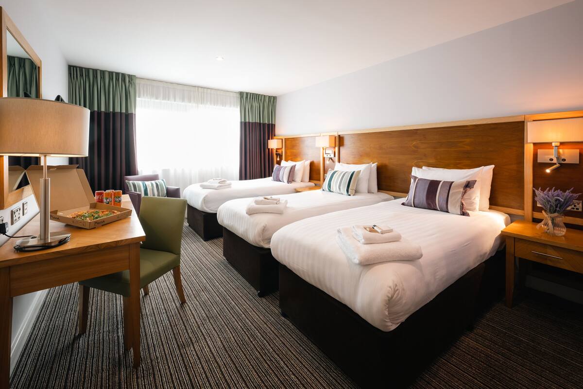 Une chambre d’hôtel avec deux lits, un bureau avec une lampe, une chaise et une petite table sur laquelle se trouve une boîte à pizza.
