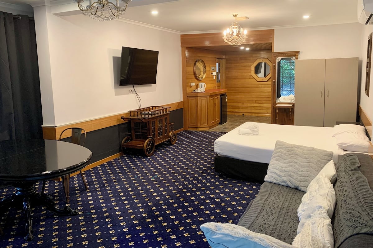 A hotel room with a bed, a sofa, a desk with a television, a wardrobe, and a chandelier.