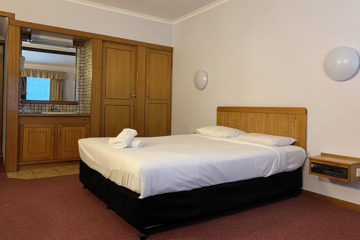 A hotel room with a bed, a wooden wardrobe, a mirror, a wall-mounted light, and a small wooden shelf.