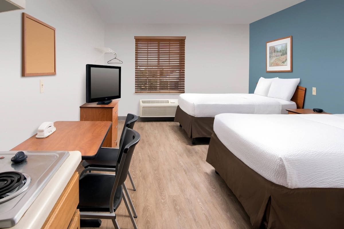Ein Hotelzimmer mit einem Bett, einem Schreibtisch mit Computer, einem Sessel, einem Fernseher und einem Fenster mit Jalousien.