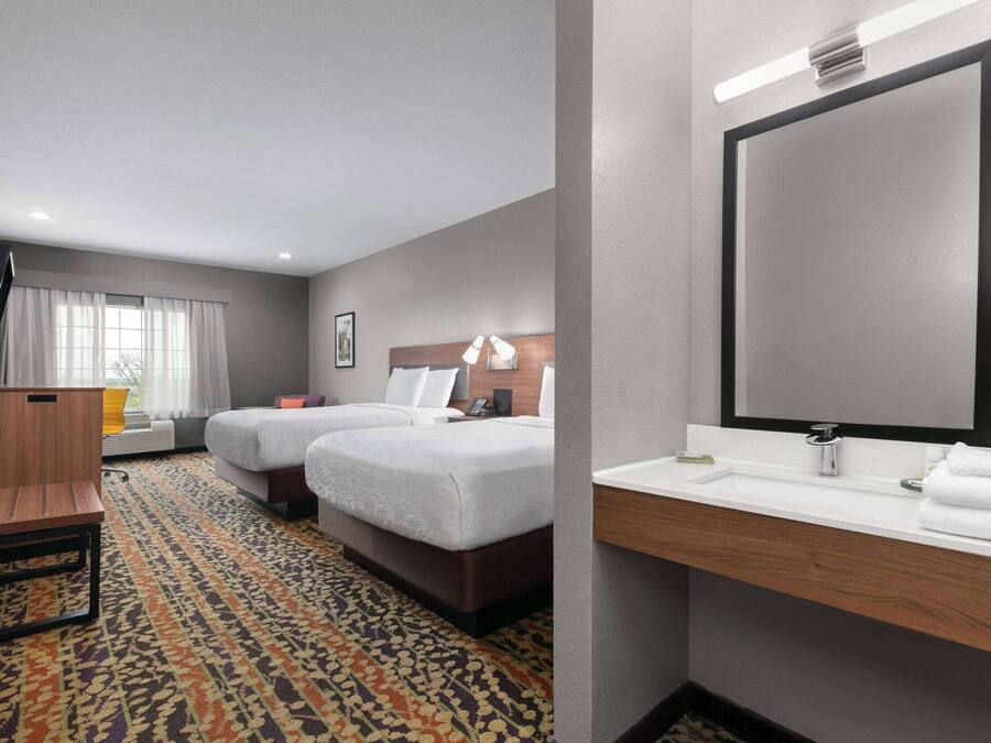 A hotel room with two beds, a desk, a large mirror, and a bathroom with a sink and towel rack.