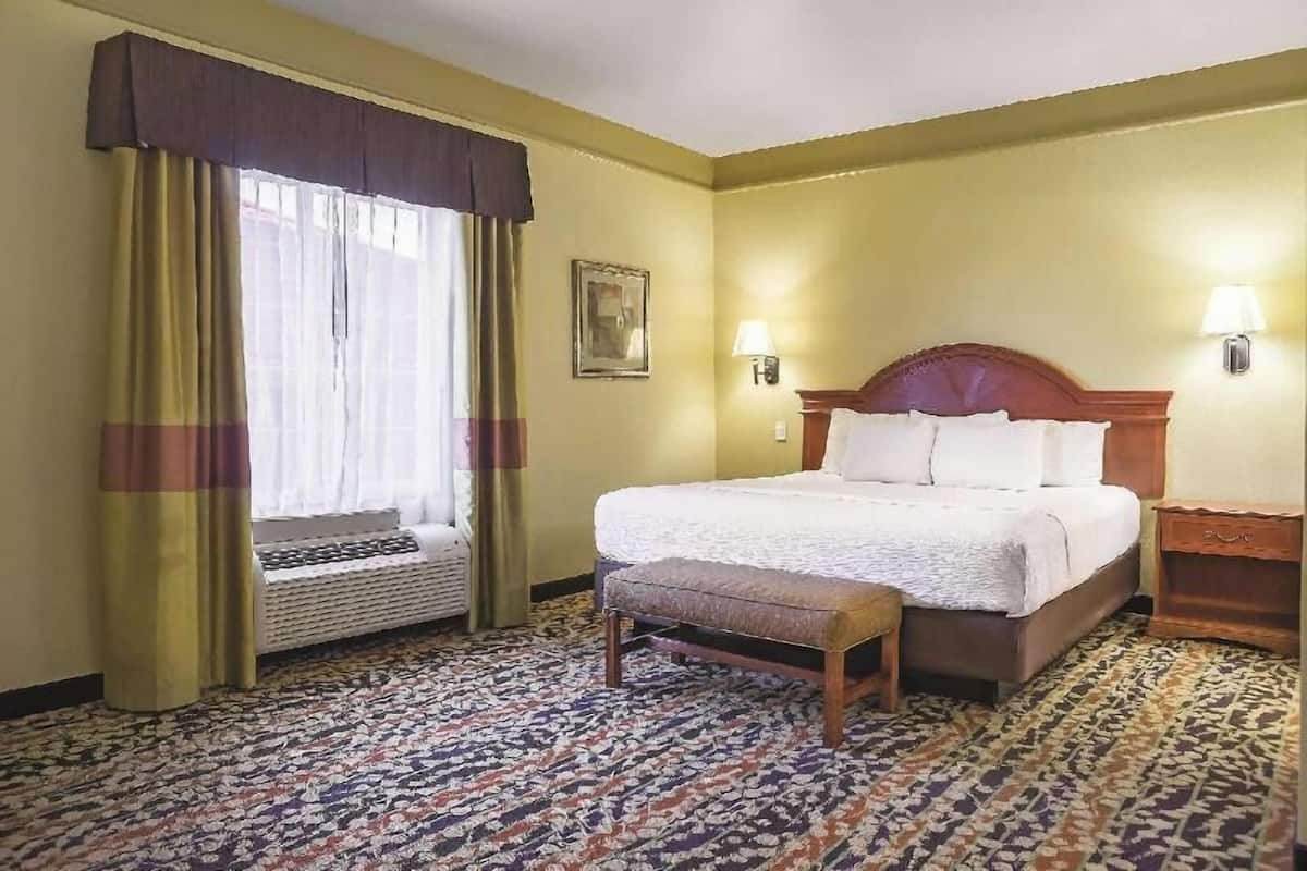 A hotel room with a bed, a bench, a window with curtains, a wall-mounted lamp, and a patterned carpet.