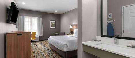 Habitación de hotel con cama, escritorio, silla, televisor y baño con lavamanos y espejo.