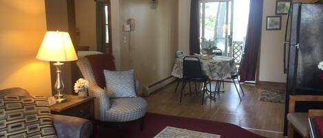 Uma sala de estar com sofá, poltrona estampada, mesa de jantar com cadeiras, geladeira e luminária.