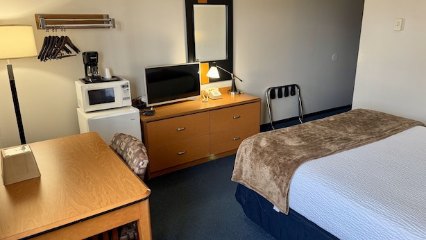 A hotel room with a bed, a desk with a lamp, a microwave, a coffee maker, a dresser, and a chair.