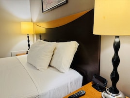 Una habitación de hotel con una cama, almohadas, un control remoto, una lámpara y un cuadro colgado en la pared.