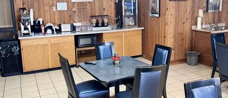 Cocina compacta con microondas, cafetera y refrigerador pequeño. Hay una zona de comedor con mesa y sillas.