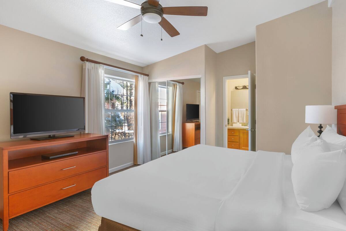 A hotel room with a bed, a TV on a wooden dresser, a ceiling fan, and a view of the outside.