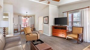 Ein geräumiges Wohnzimmer mit Deckenventilator, Fernseher auf Holzständer, Sofa, Schaukelstuhl und Essbereich mit Stühlen und Tisch.