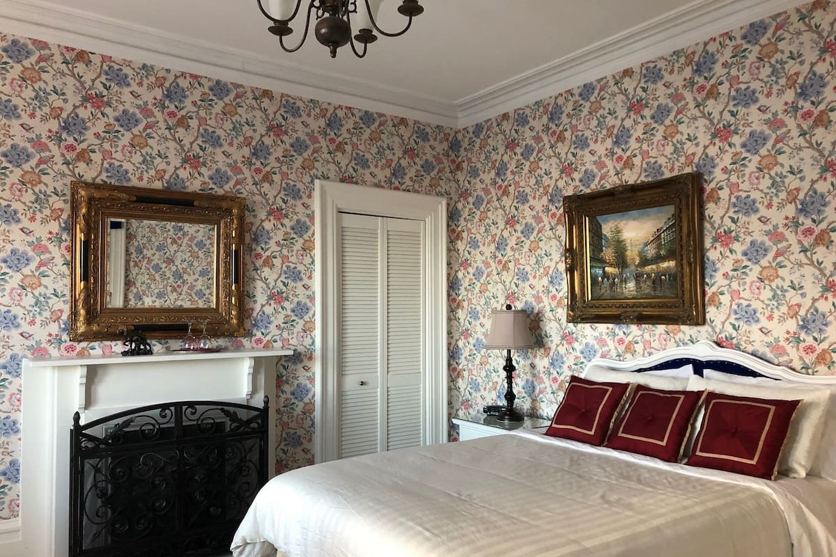 A bedroom with floral wallpaper, a fireplace, a bed with red and blue pillows, and two framed pictures on the wall.