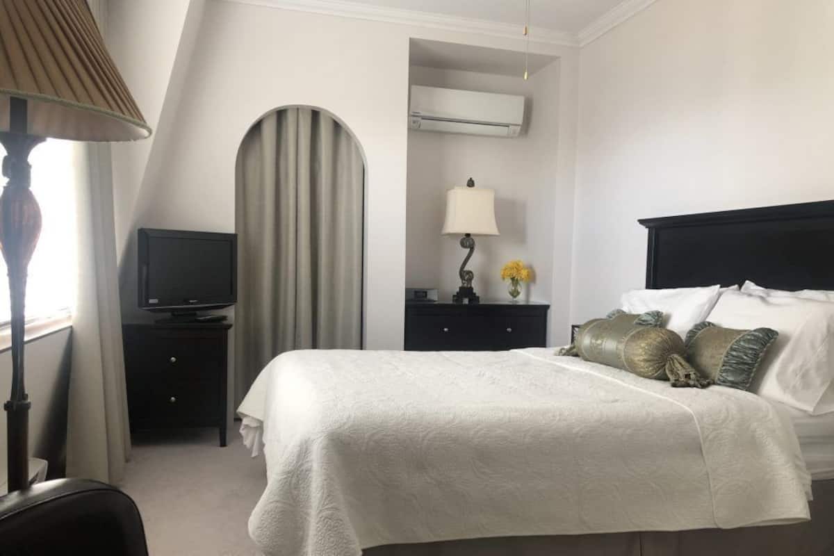 A hotel room with a bed, a television on a stand, a lamp, a dresser, and an air conditioning unit.