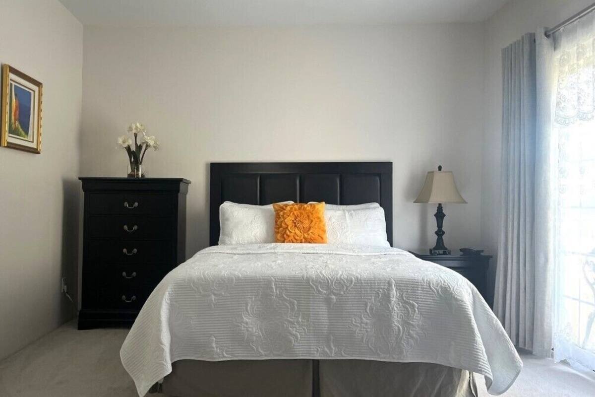 A neatly arranged bedroom with a bed, a dresser, a lamp, and a window with curtains.