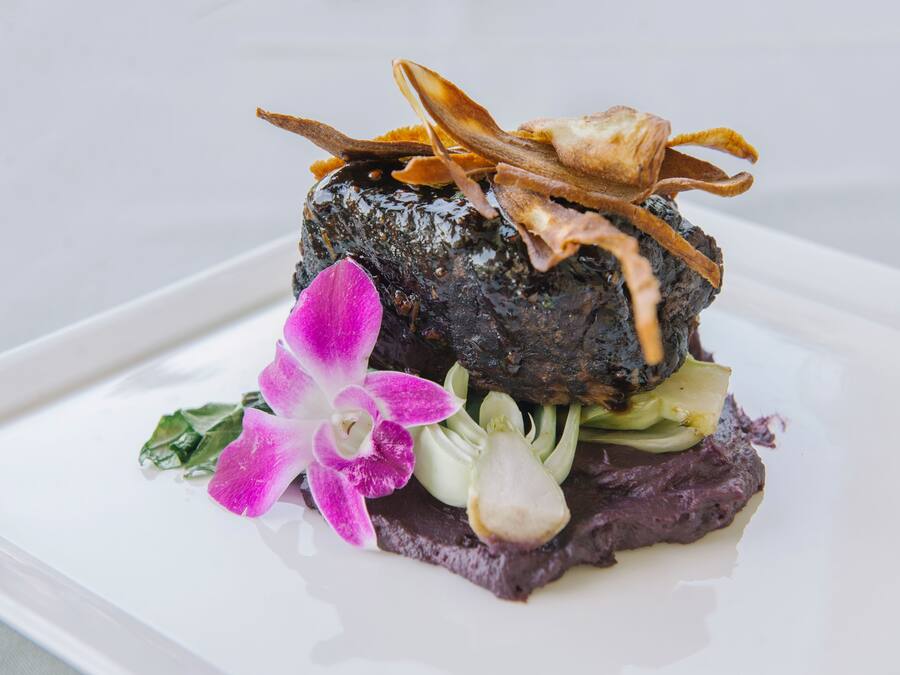 A dish with a dark, textured base, garnished with thinly sliced meat and a flower.