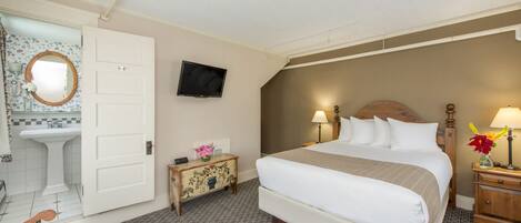 A hotel room with a bed, a television, a bedside table with a lamp, a nightstand with a vase of flowers, and a bathroom with a sink and mirror.