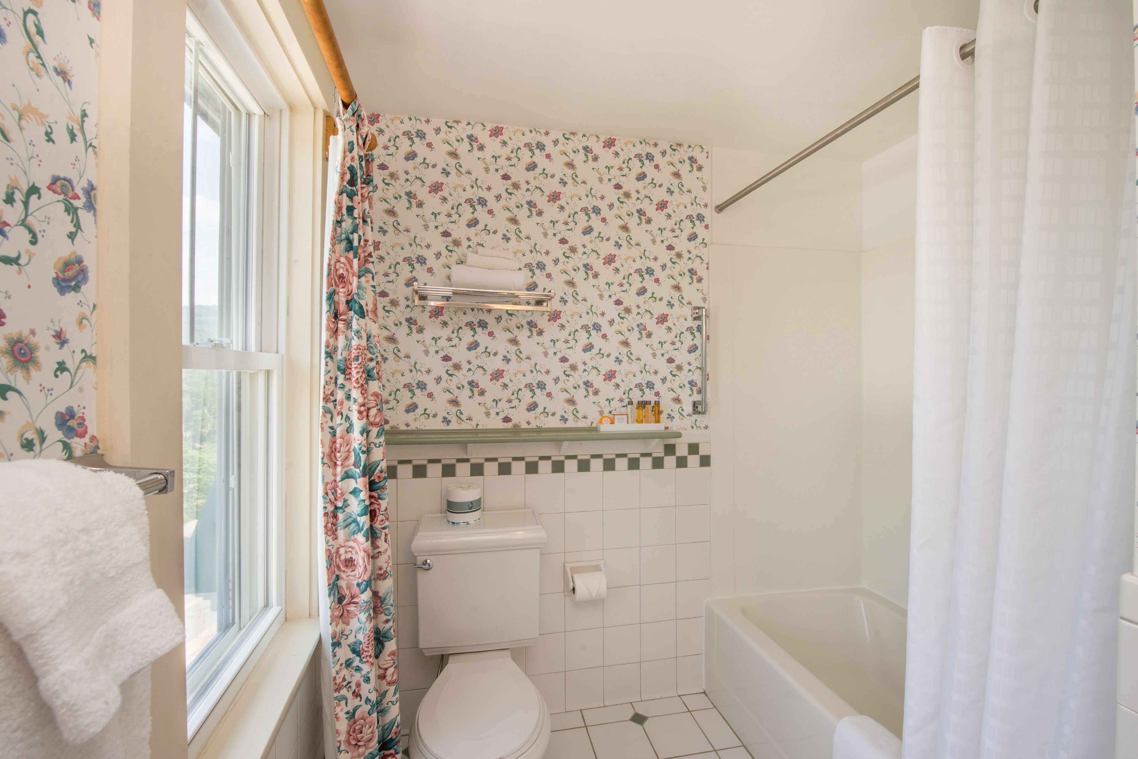 A bathroom with a toilet, shower, and a window with a floral curtain.