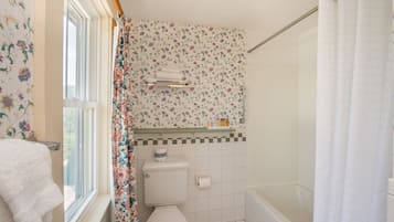 A bathroom with a toilet, shower, and a window with a floral curtain.