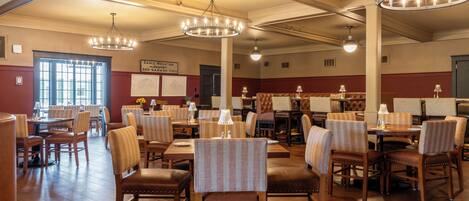 A spacious dining area with multiple tables and chairs, high ceilings with exposed beams, and a large window providing natural light.