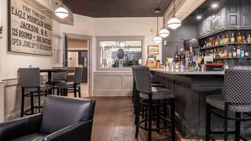 A bar area with a bar counter, stools, and a variety of bottles. There is a sign on the wall and a window in the background.